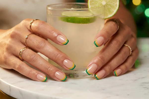 Close-up de mãos femininas elegantes exibindo tendências de unhas decoradas para a copa 2026. A nail art traz um design moderno e seguro no estilo "Brazilcore", com base nude e francesinha dupla em verde e amarelo. Destaca a importância dos cuidados com as unhas e de uma esmaltação perfeita feita com segurança.